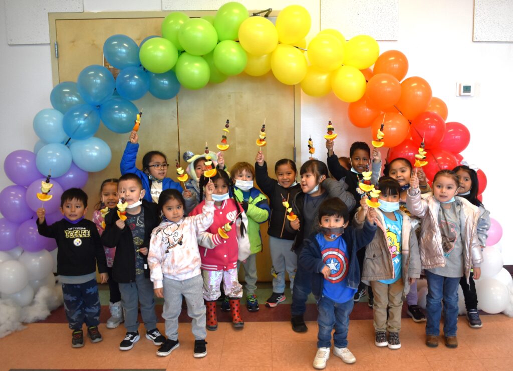 Kids hold fruit under a rainbow balloon arch