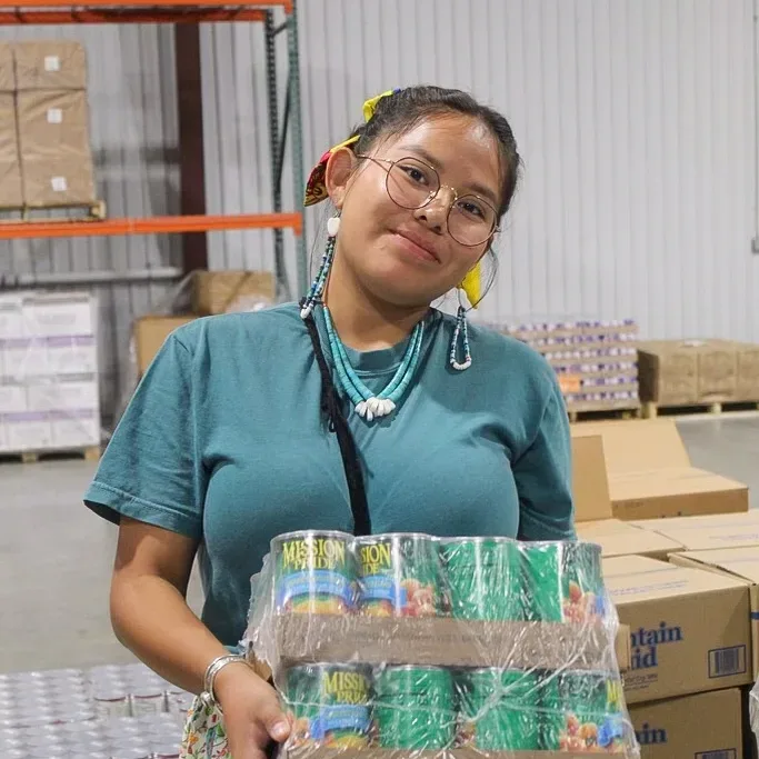 A young person with a braid holds cases of canned foods