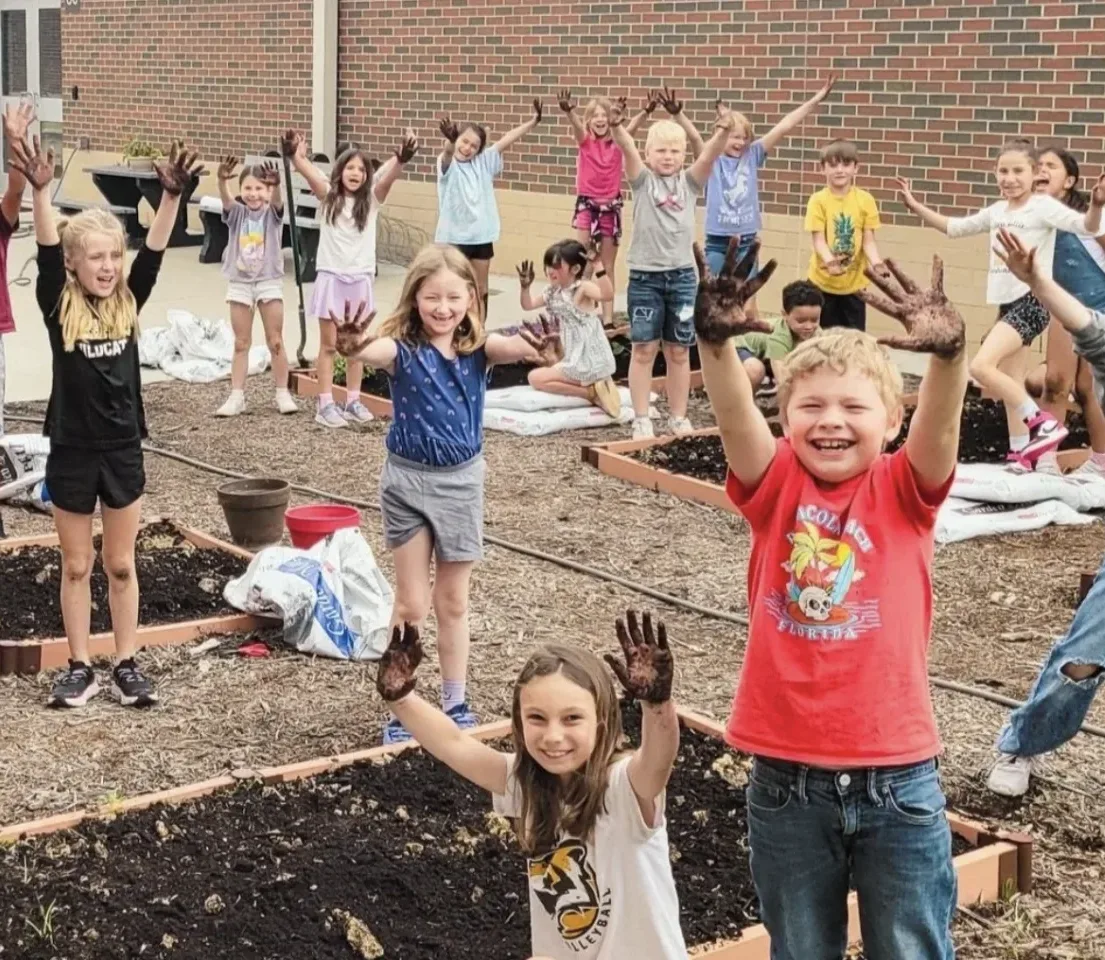 Kids stand excitedly around garden beds with dirt on their hands